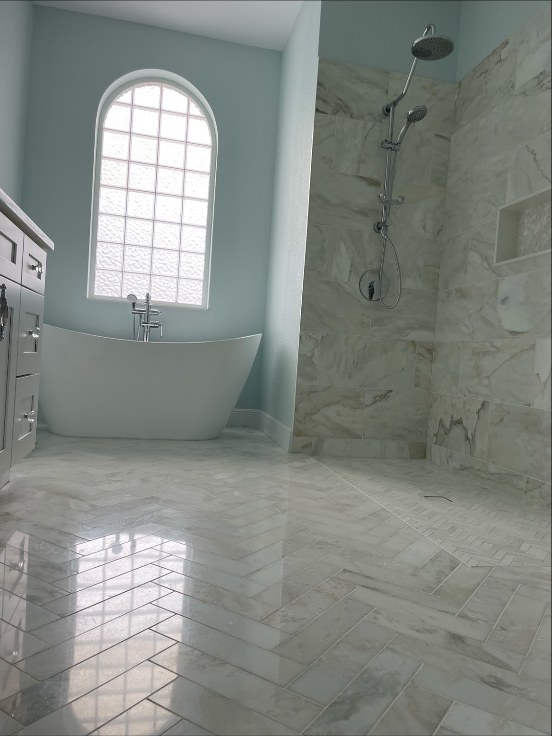 A finished bathroom remodel by Best Services of Remodeling: a freestanding soaking tub beneath a tall arched window with marble tile walls and a herringbone marble floor.